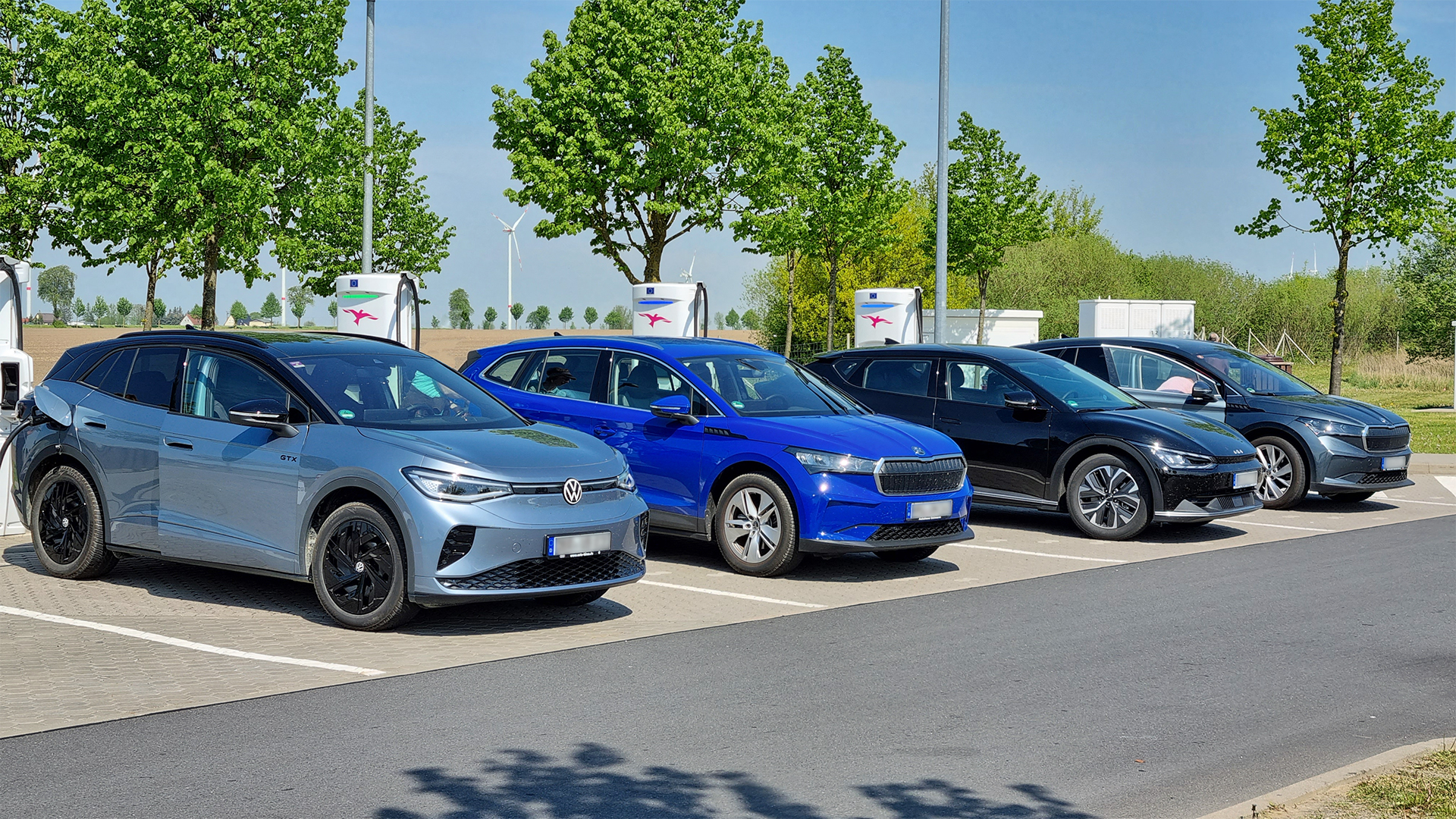 Quatre cotxes elèctrics de diferents fabricants alineats en un hub de càrrega ràpida al costat d'una autopista.